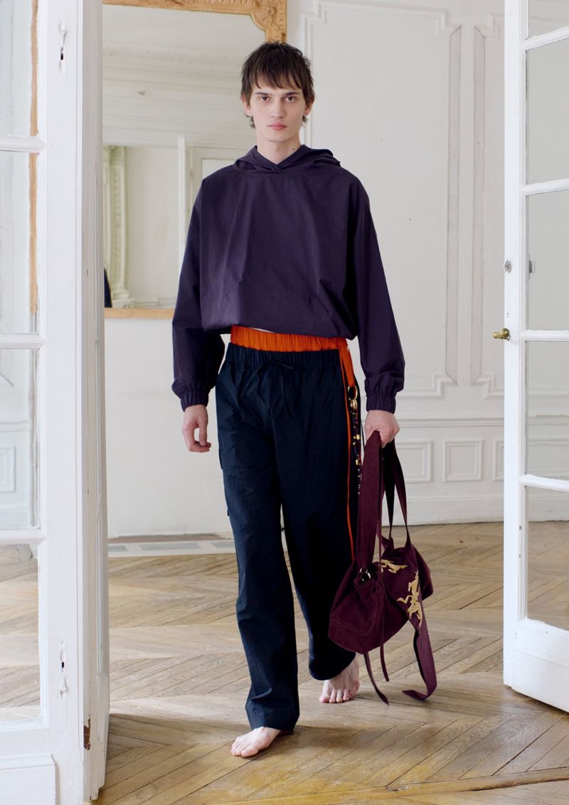 Young adult man models dark purple hoodie and dark pants, carrying a drawstring bag in a brightly lit room with white walls.