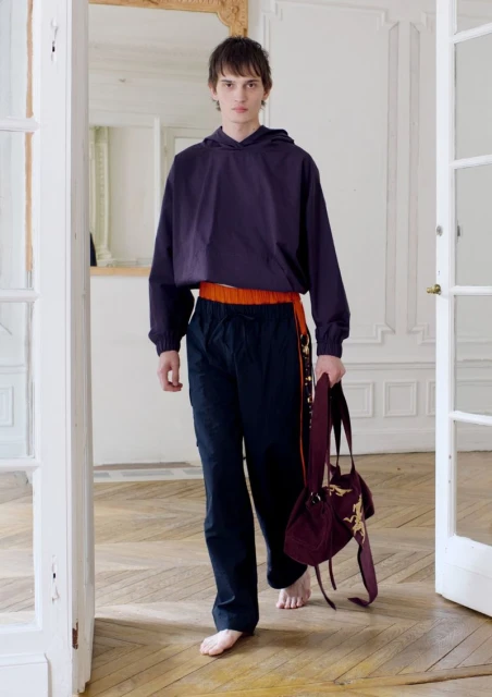 Young adult man models dark purple hoodie and dark pants, carrying a drawstring bag in a brightly lit room with white walls.