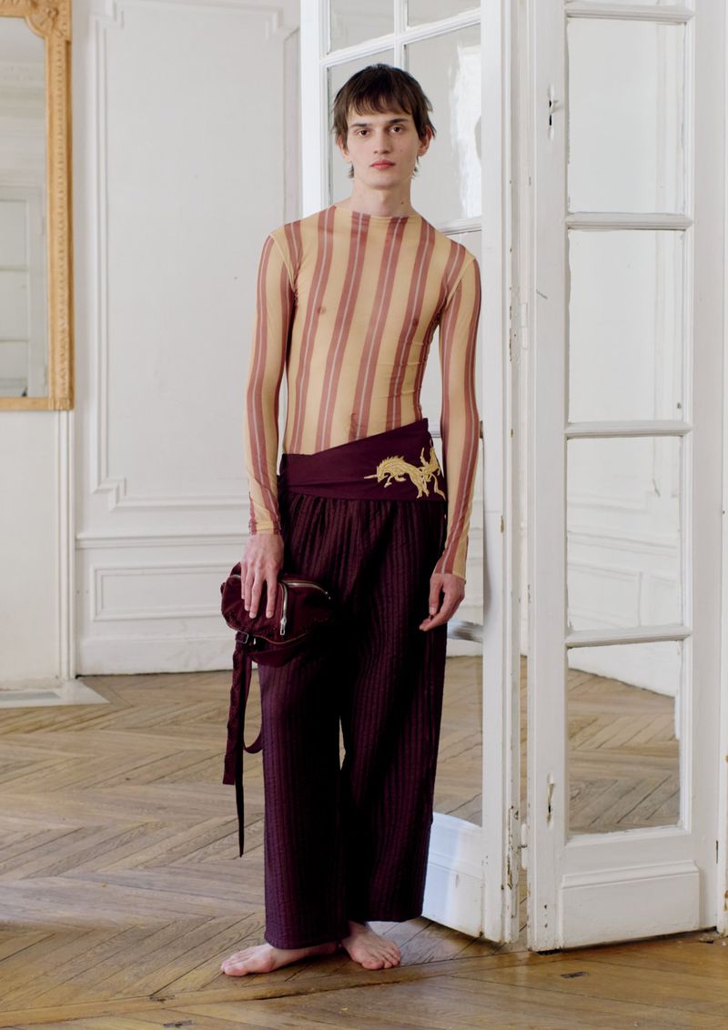 A young adult male poses in a sheer striped shirt and maroon pants with a matching purse in an indoor fashion setting with white walls.