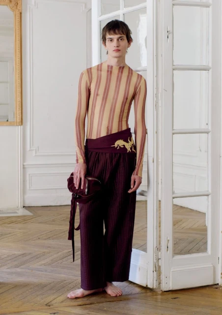 A young adult male poses in a sheer striped shirt and maroon pants with a matching purse in an indoor fashion setting with white walls.