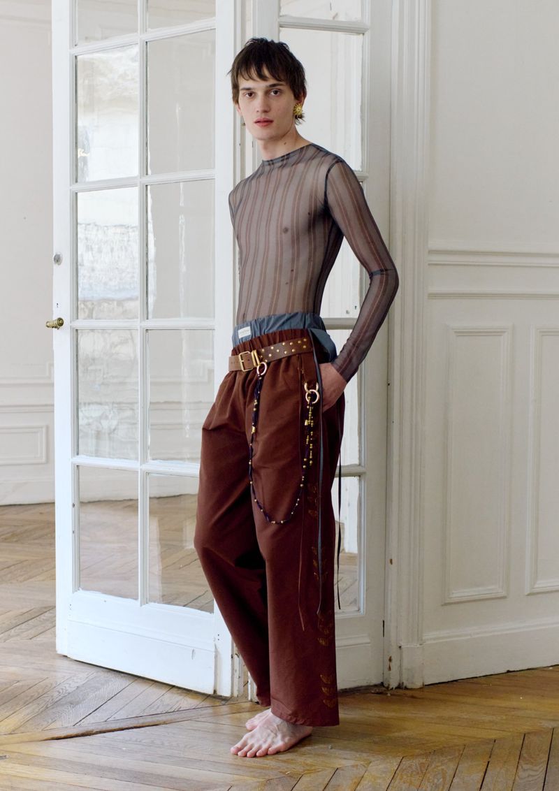 Fashionable young adult with dark hair and sheer top poses near white-framed glass doors with brown trousers and gold chain belt detail.