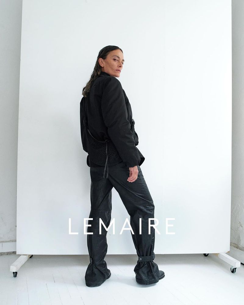 Adult female model wearing black jacket and black pants in front of a white screen in a photo studio looks back at the camera.