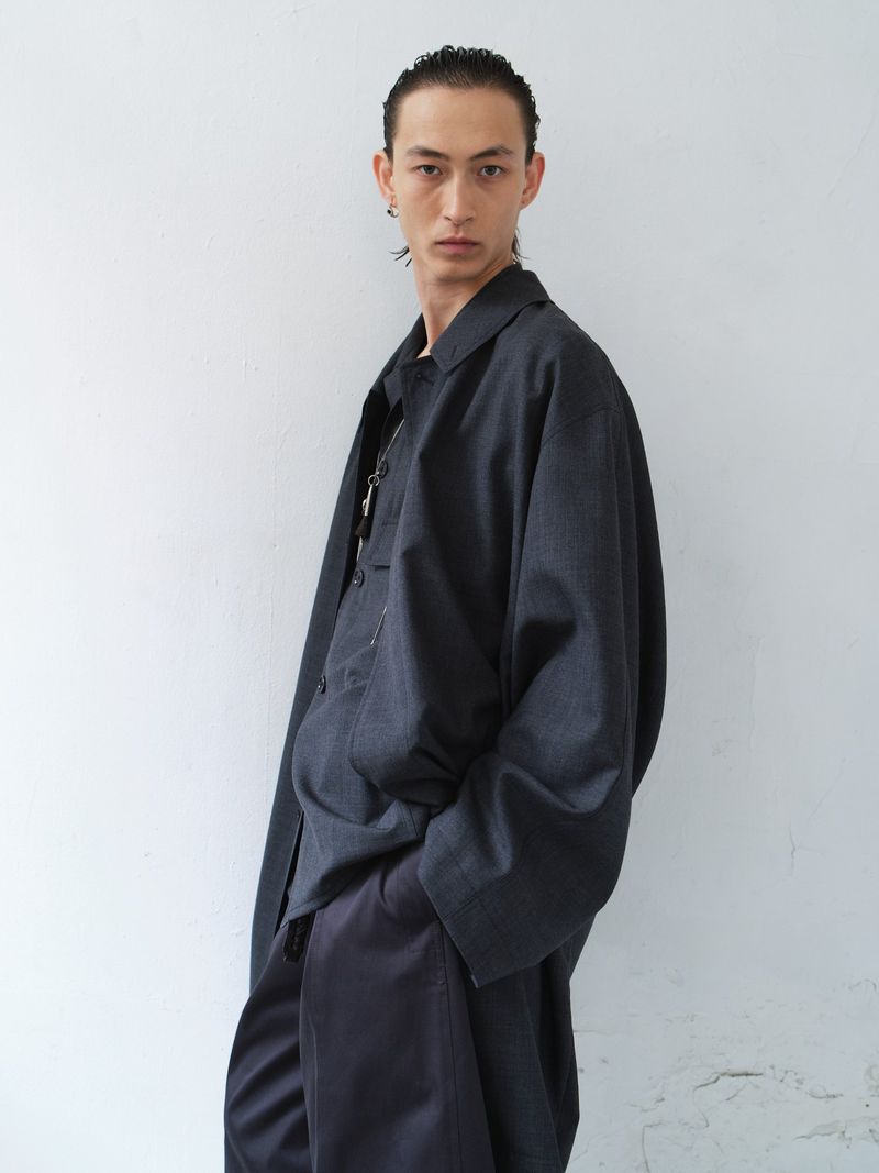 An adult male poses in front of a light grey wall, wearing a dark button-down shirt and pants in a medium, portrait-oriented shot.