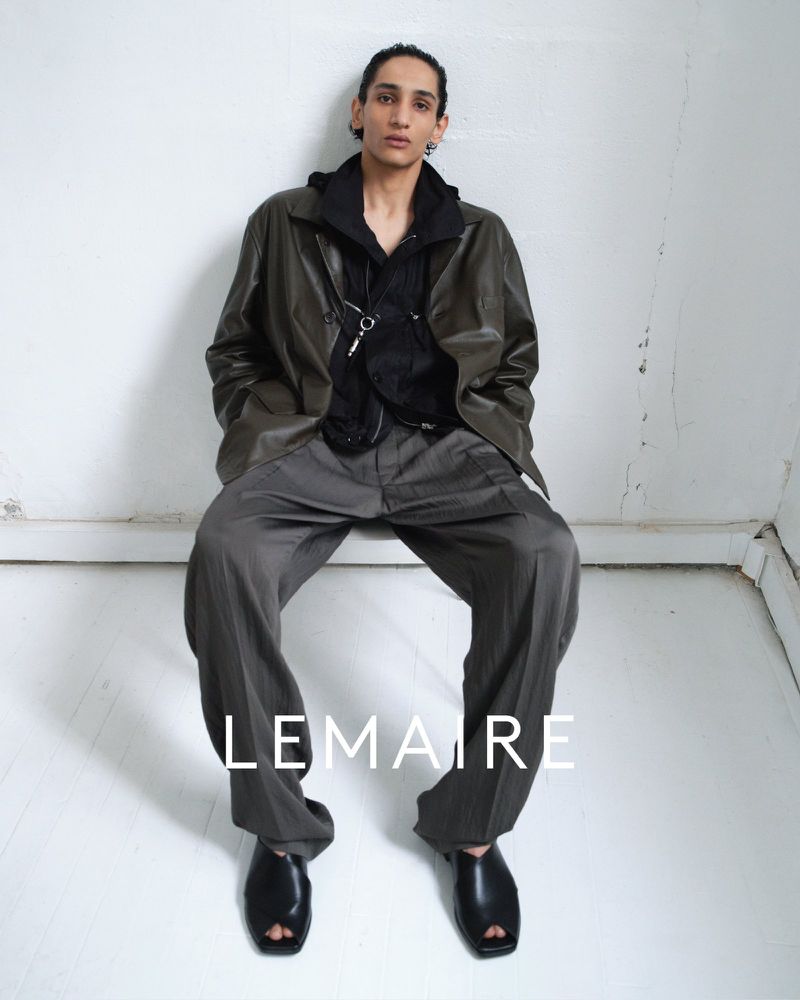 Fashion shot of a model sitting in a studio, wearing a hooded shirt, jacket, wide pants, and sandals, with the brand name LEMAIRE visible.