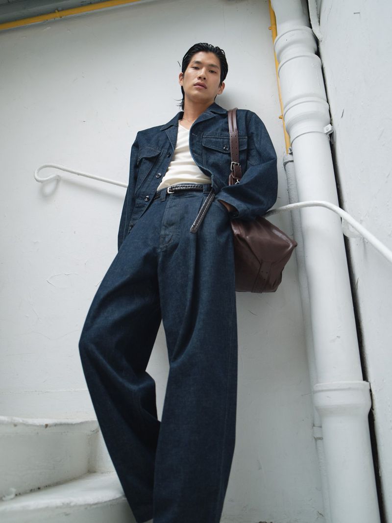 Man in denim jacket and wide-leg jeans poses against white wall with brown leather messenger bag in a low-angle, full shot.