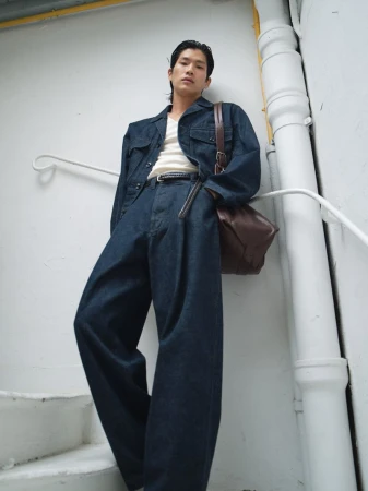 Man in denim jacket and wide-leg jeans poses against white wall with brown leather messenger bag in a low-angle, full shot.