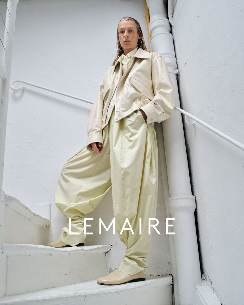 Fashion shot of woman in white jacket and trousers, posing on stairs in bright light. The brand, LEMAIRE, is visible in the image.