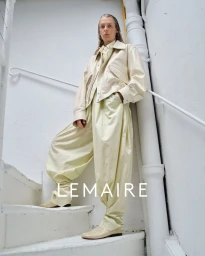 Model standing on a white staircase wearing a cream leather jacket and pale yellow trousers for Lemaire Spring-Summer 2025.