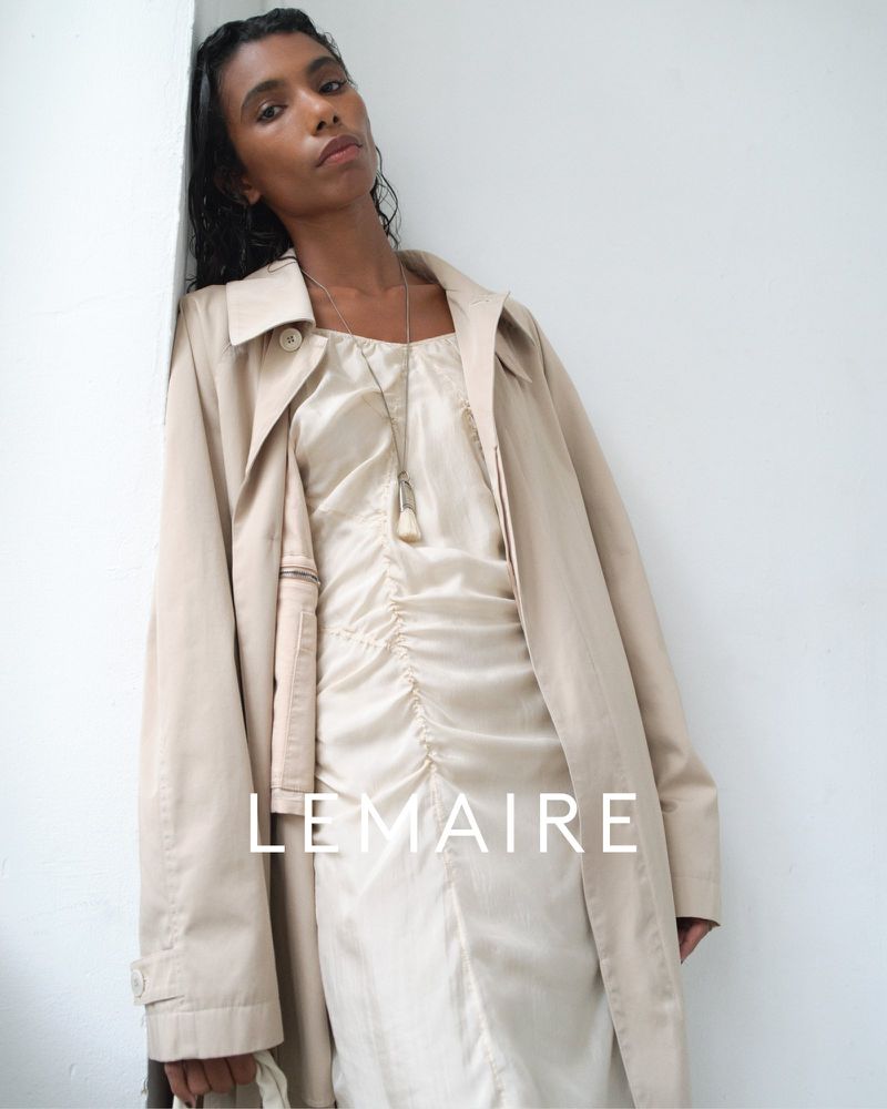 Model leaning against a white wall wearing a beige trench coat over a cream ruched dress for Lemaire spring summer 2025.