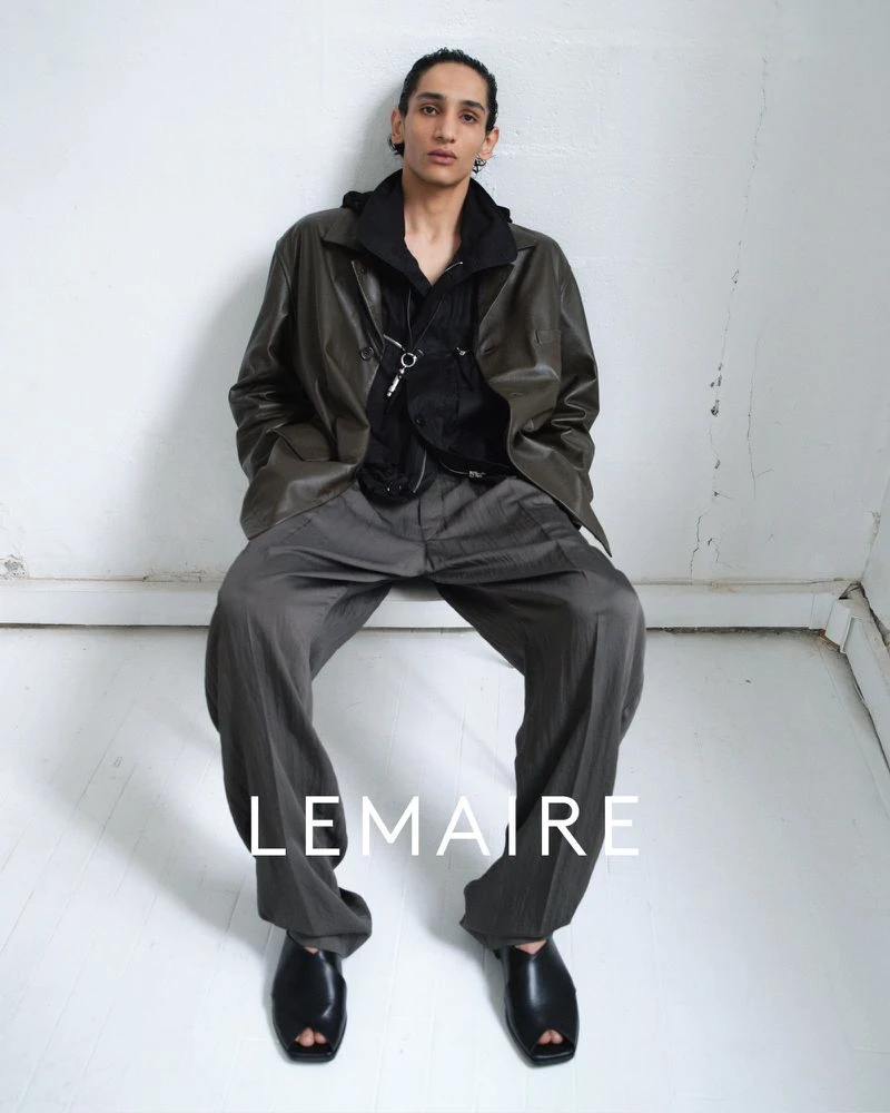 Model seated against a plain white wall wearing an olive leather jacket, black hoodie, and grey trousers.
