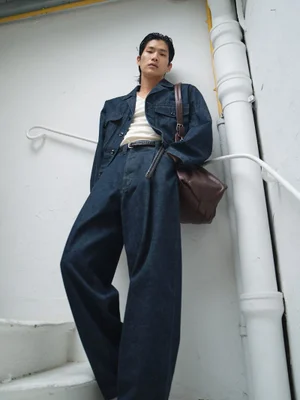 Man wearing a dark denim jacket and matching trousers leaning against a white stairwell wall holding a brown leather shoulder bag.