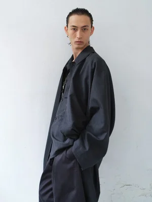 Model in dark charcoal button-down shirt and matching coat standing against a pale white wall