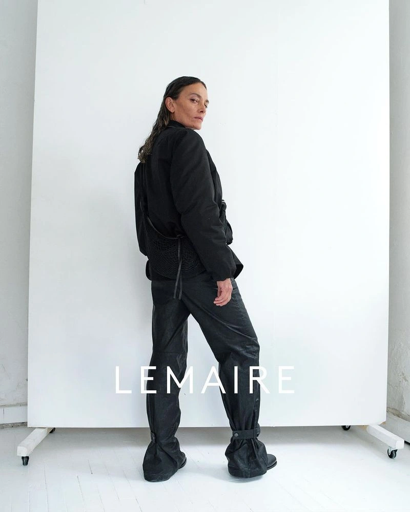Model wearing a black jacket and dark trousers standing against a white seamless backdrop for Lemaire Spring-Summer 2025.