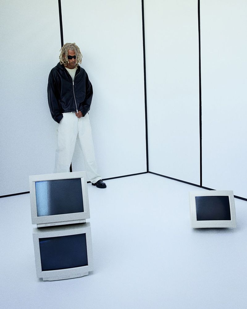 Man in black jacket and white pants poses in a white studio with stacked vintage computer monitors and single monitor. 