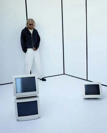 Man in black jacket and white pants poses in a white studio with stacked vintage computer monitors and single monitor. 