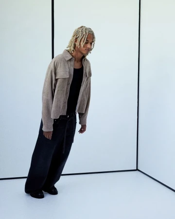 Stylish adult male with blond dreadlocks in an oatmeal-colored shirt and dark jeans poses in a bright photo studio with white walls.