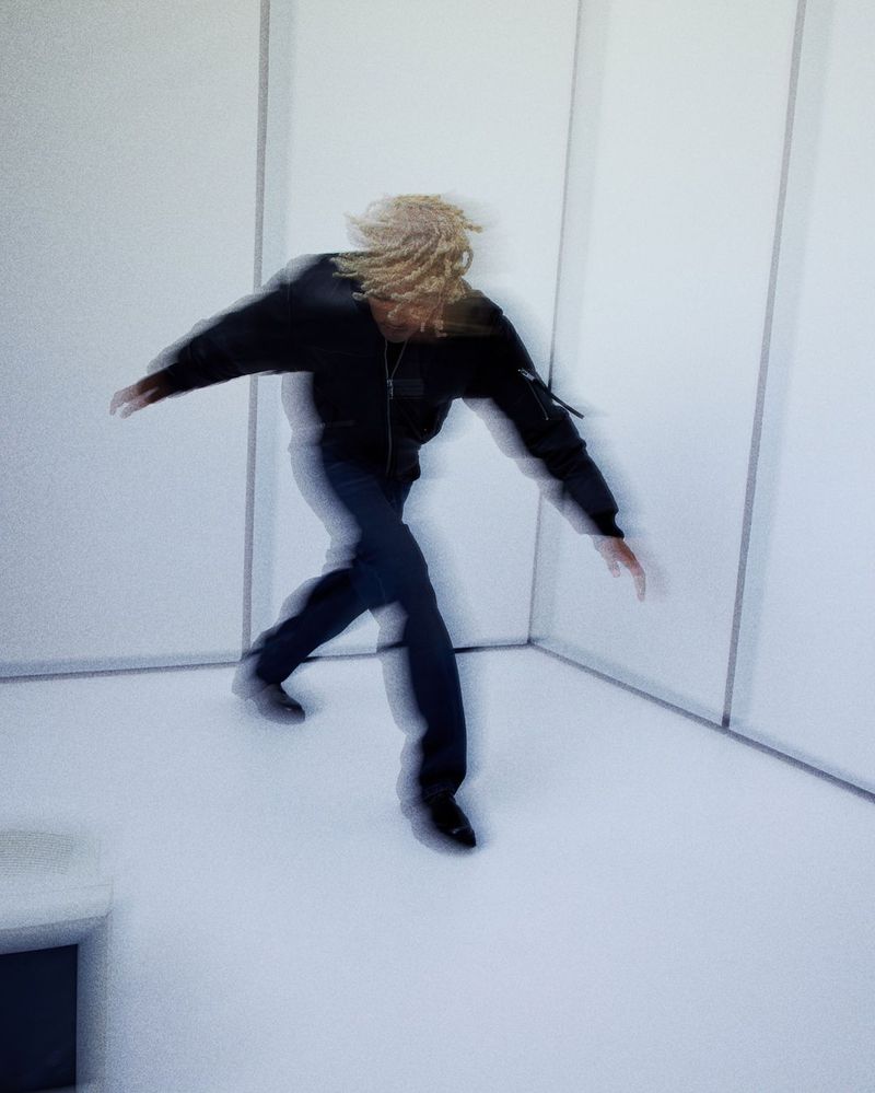 Blurred image of an adult with blonde dreadlocks wearing a black jacket and blue jeans, posing indoors against a white wall.