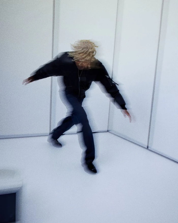 Motion blur photograph of a man wearing a black leather jacket and dark trousers inside a minimalist white room