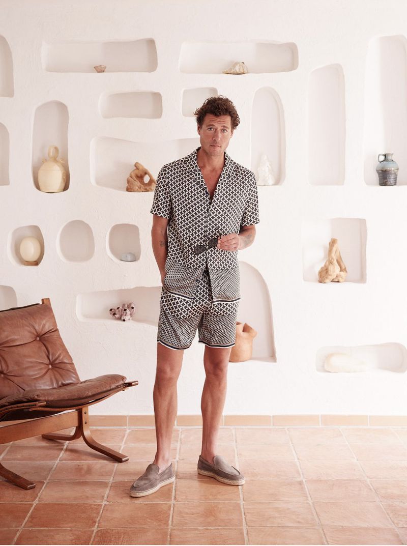 A stylish adult male in a patterned shirt and shorts poses in an indoor setting with terracotta floors and white wall decor.
