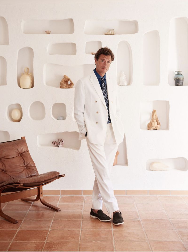Man in a white suit posing indoors with chair and decorative wall, creating a sophisticated and classic portrait.