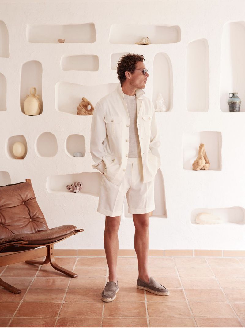 A stylish adult male in a white linen outfit poses in front of a textured white wall with niches, complemented by a brown leather chair.