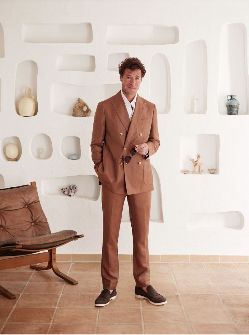 A full shot of a man in a brown suit, standing indoors with a hand in his pocket, wearing dark shoes, with art objects on a white wall.