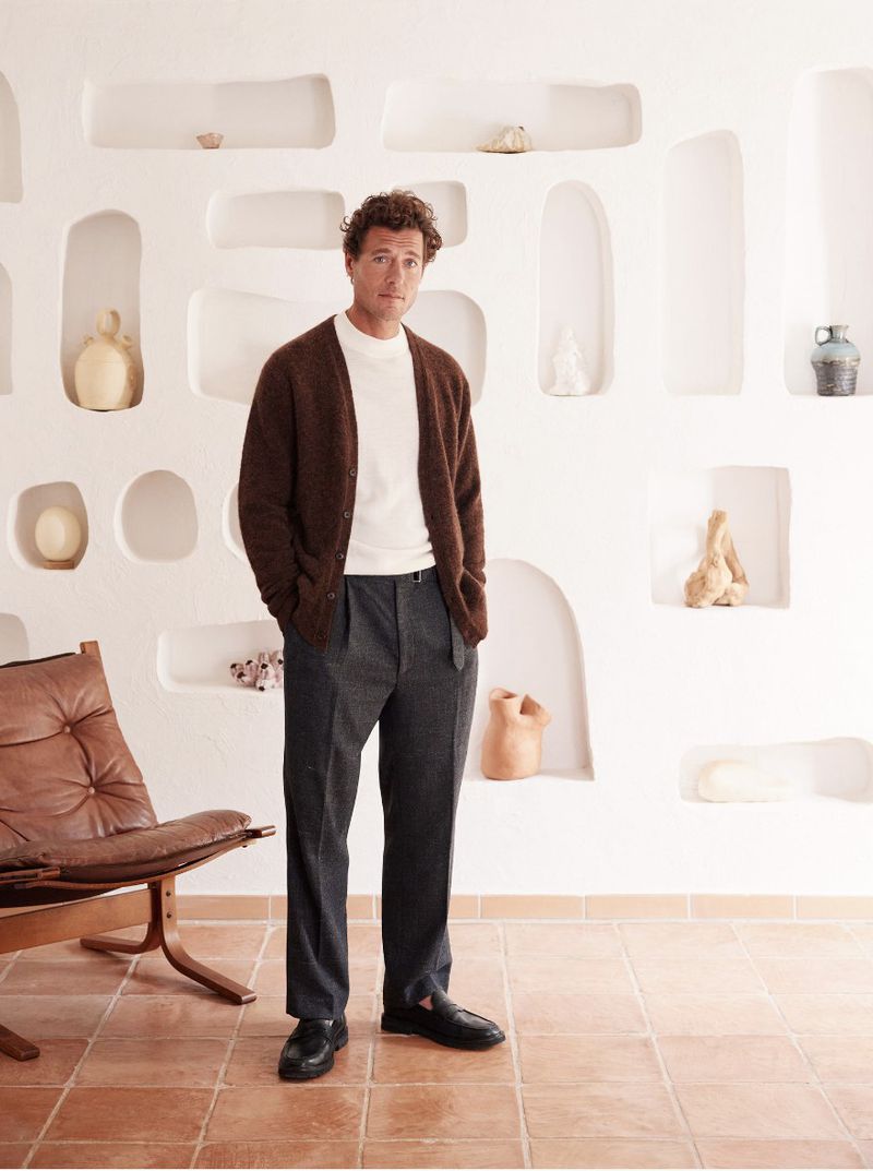 Full body portrait of a man in a cardigan and slacks standing against a white wall with niches, inside a minimally furnished room.