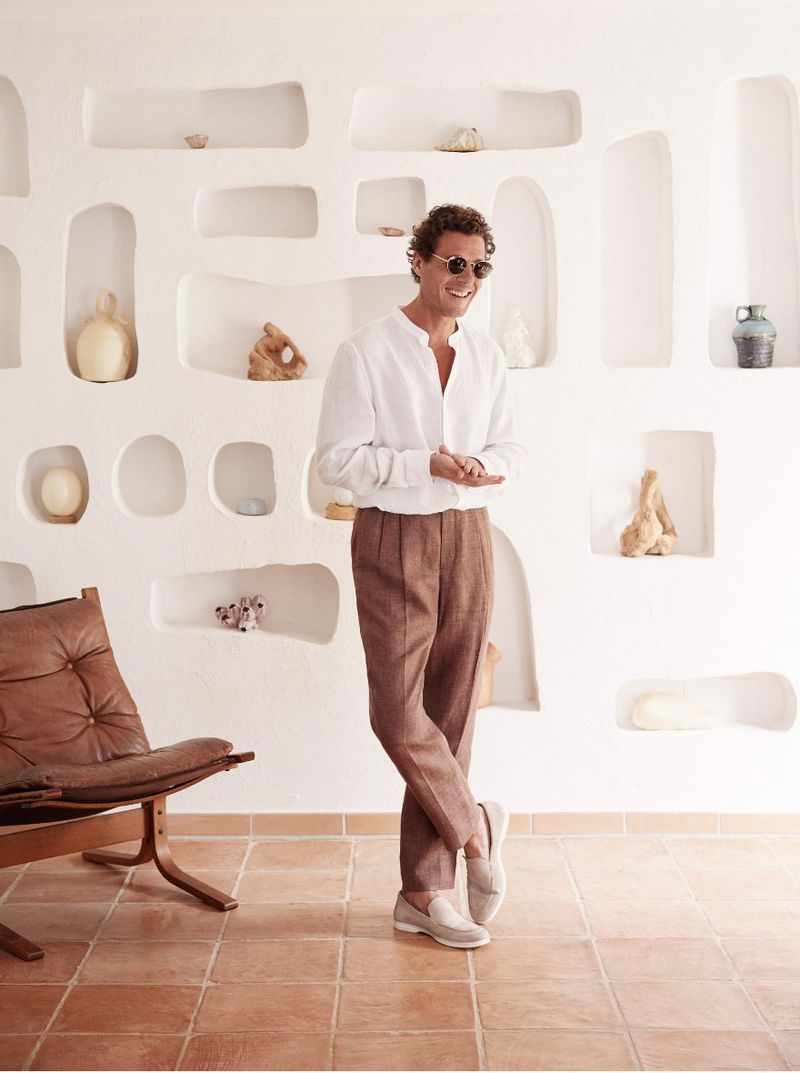A man in a white shirt and brown pants stands indoors next to a brown leather chair, with a wall of display niches in the background.