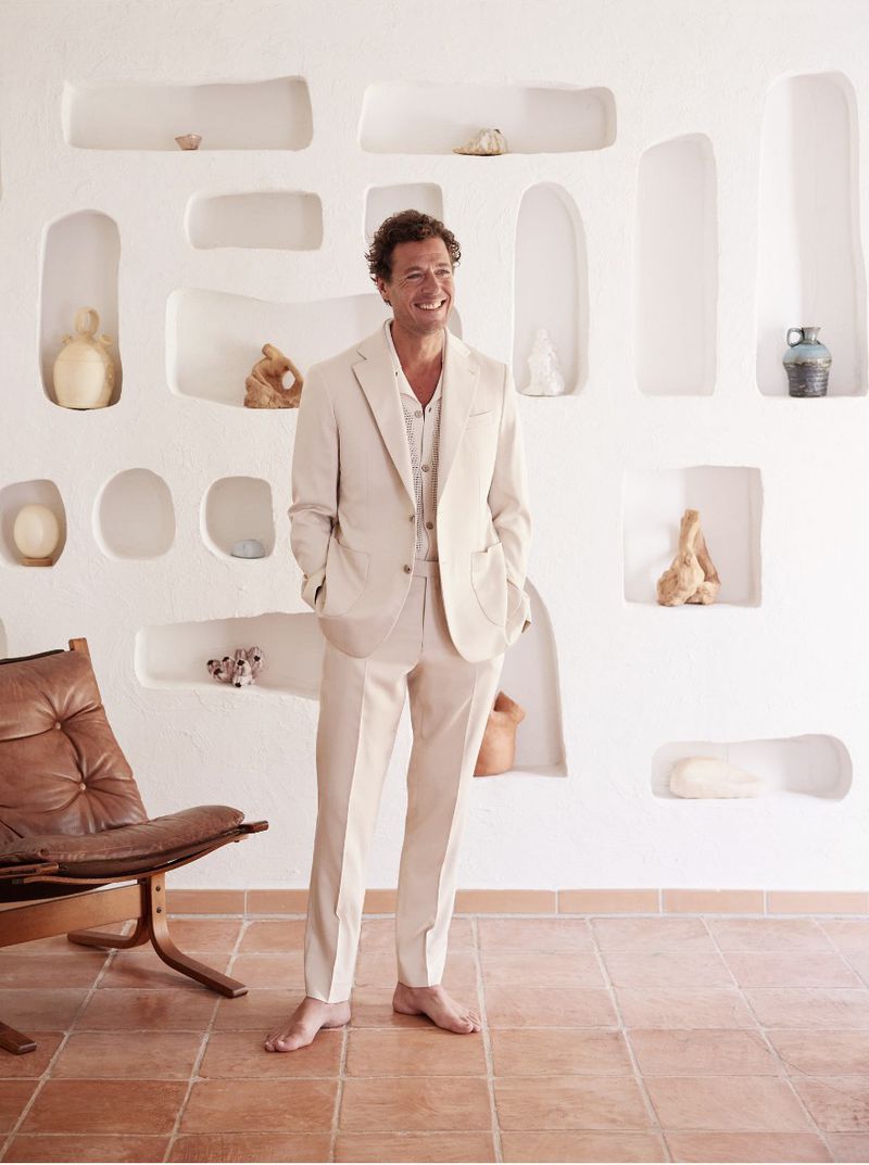 Smiling adult man in a beige suit stands barefoot in a room with light walls, inset shelves, a leather chair, and terracotta tile floors.