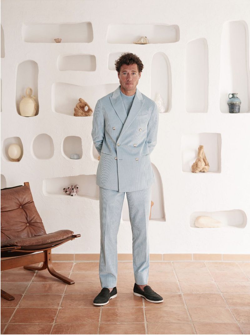Full-length shot of a man in a light blue suit posing indoors against a decorative white wall, complemented by a leather chair and tiled floor.