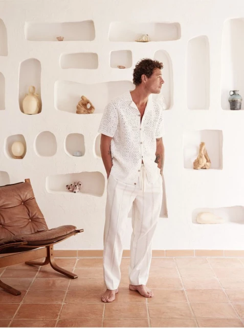 Man in a white eyelet shirt and striped pants stands barefoot in an indoor setting with a light-filled minimalist design.