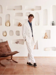 Man wearing a white double-breasted suit standing against a textured white wall with inset wall niches.