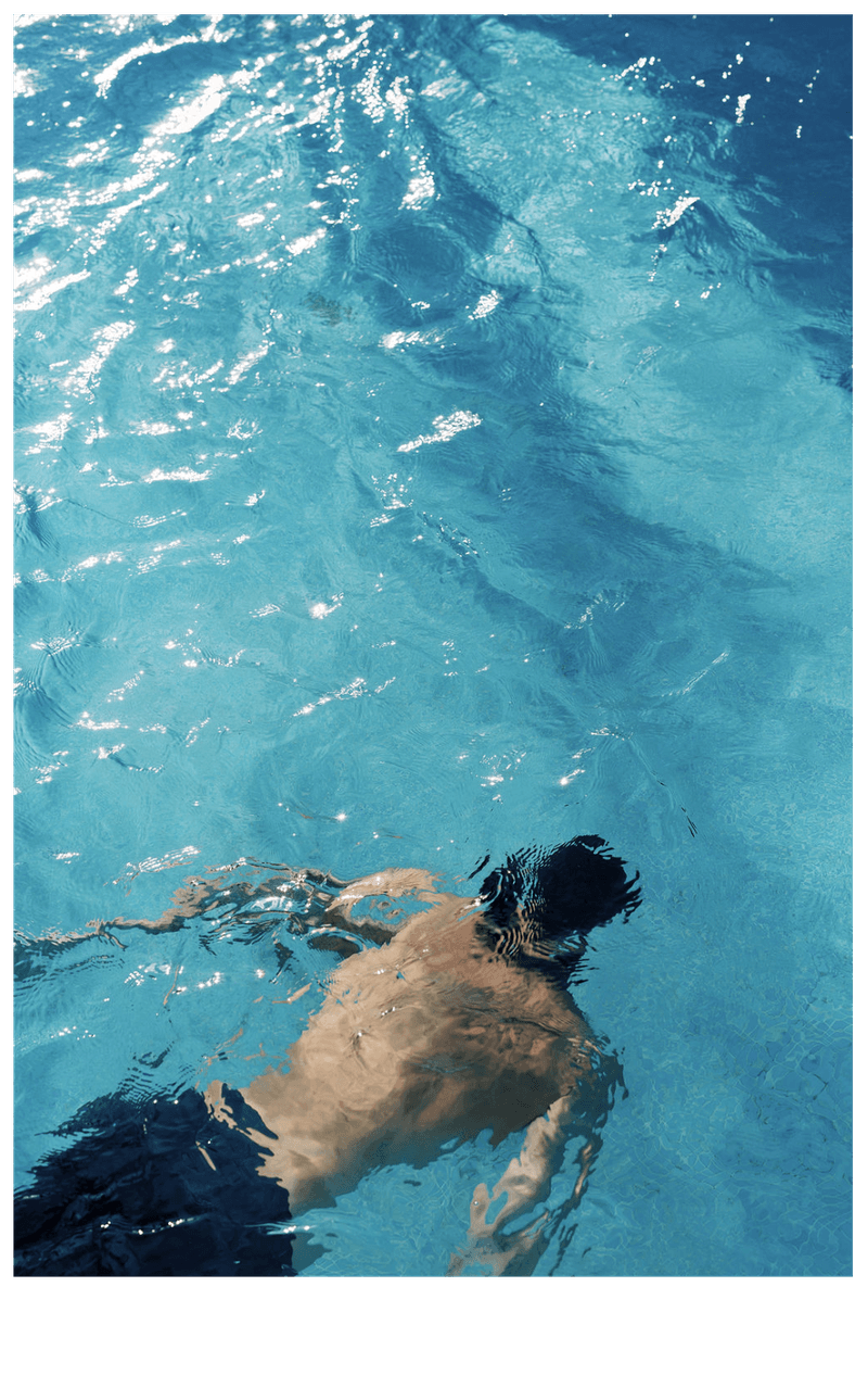 Adult swimming in clear, light-blue water, captured from an overhead angle displaying only the back and head above water.