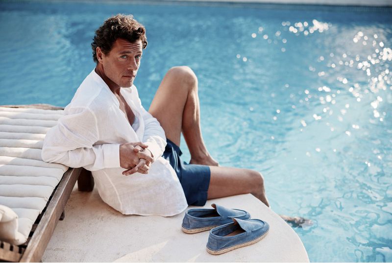 Adult male poses outdoors in the sunshine, wearing a linen shirt and shorts, near a pool, with a pair of shoes in the foreground.