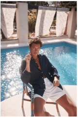 Man in dark shirt and white shorts seated on a chair by a swimming pool for Moss Resort SS24.