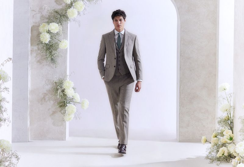 Man in a gray three-piece suit walks through an arched entryway framed by white flowers. Fashion shot with shallow depth of field.