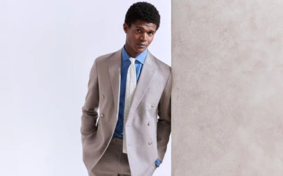 Model in a grey double-breasted suit with a blue shirt and white tie leaning against a pillar.