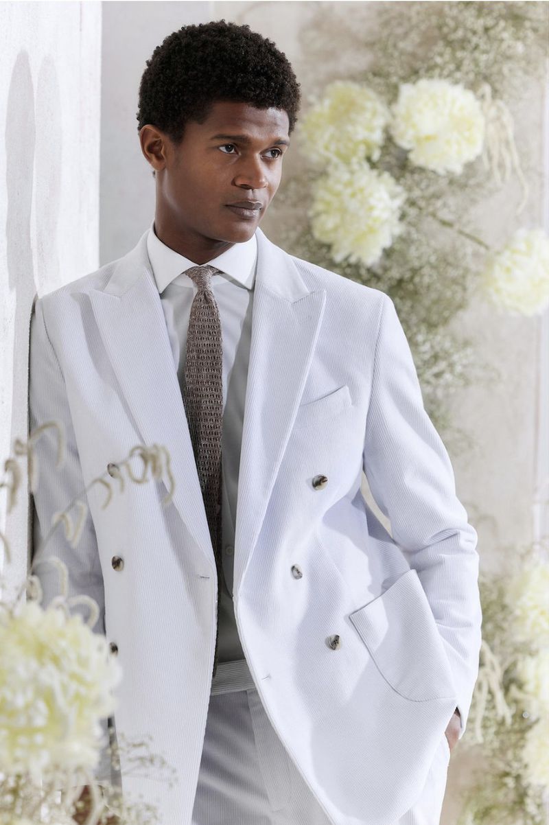 Portrait of a handsome man in a light blue suit standing against a white wall adorned with soft white floral decorations.