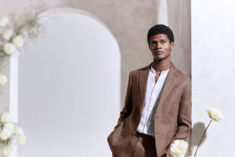 An African American man in a brown suit leans against a wall with white roses, showcasing modern fashion and elegant portraiture.