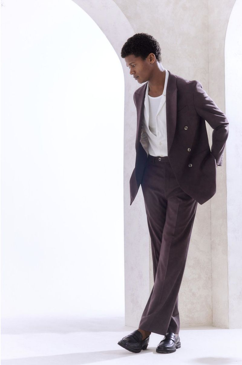 Adult male fashion model in a plum-colored double-breasted suit posing indoors against an arched architectural wall for a portrait photograph.
