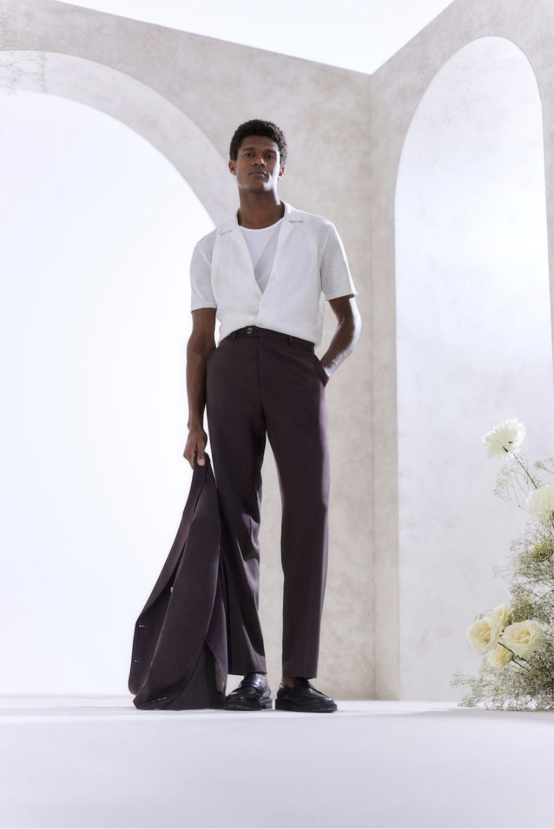 Man poses indoors wearing a short-sleeved white shirt, brown pants, and black shoes, holding a brown jacket against an off-white wall.