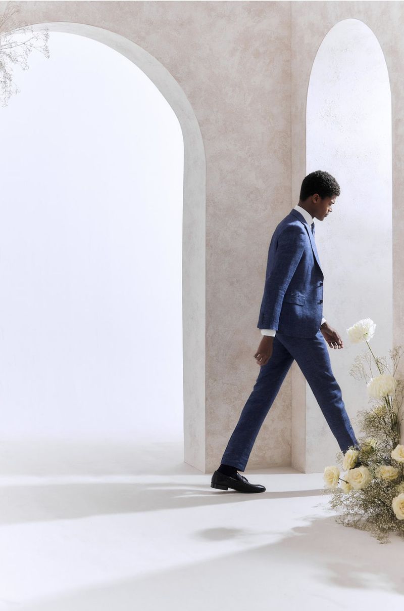 An African American man in a blue suit walks through an archway adorned with white roses in a bright, minimalist setting.