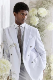 Model O'Shea Robertson in a pale blue double-breasted suit and textured tie leaning against a wall with white floral arrangements.