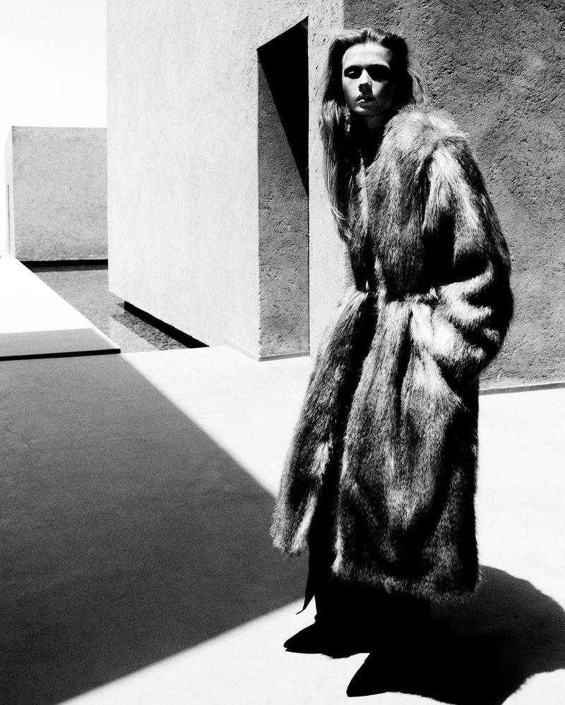Monochrome fashion shot of a person in a fur coat standing against an architectural background, strong shadows accentuate the scene.
