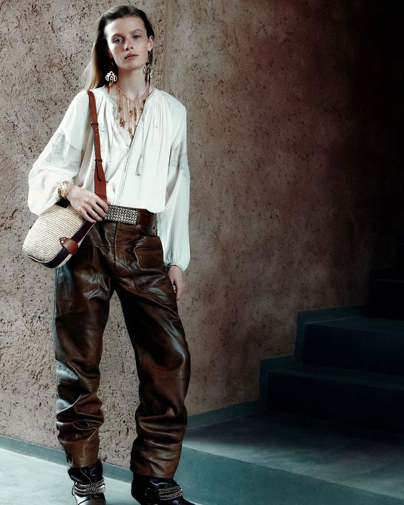 A young woman models white blouse, brown leather pants, and shoulder bag against textured wall with stairs, showcasing fashion style.