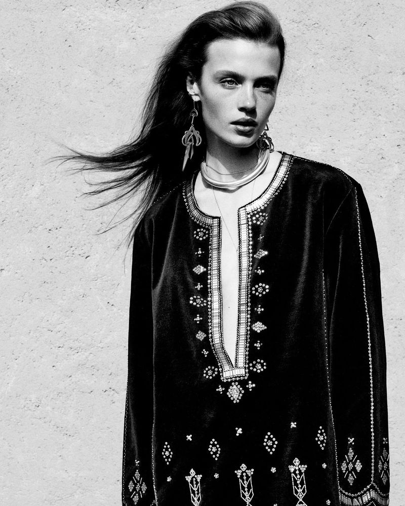 Black and white close-up of an adult woman with long dark hair wearing embellished velvet top and large earrings, against a neutral wall.