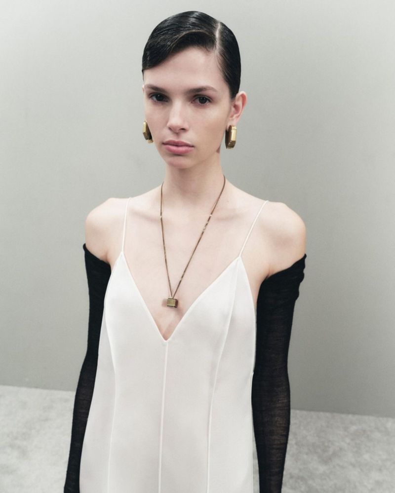 Fashion photo of a female model in a white dress and long black gloves posing in a studio with neutral background. Earring and necklace details.
