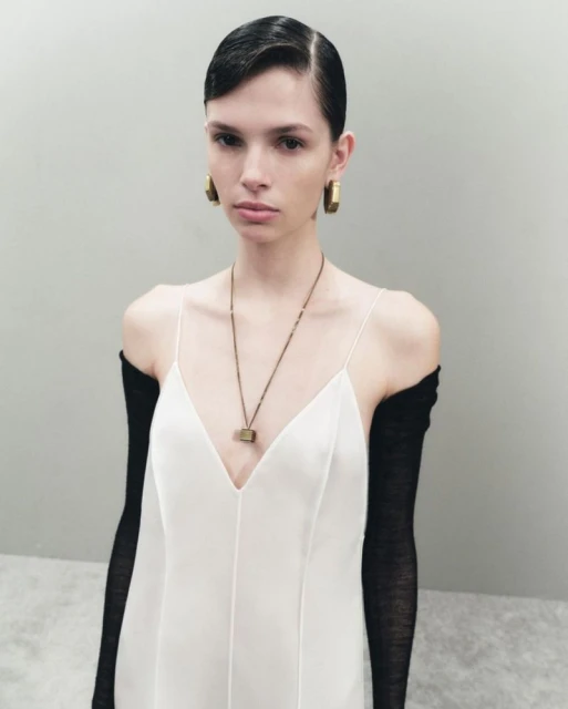 Fashion photo of a female model in a white dress and long black gloves posing in a studio with neutral background. Earring and necklace details.