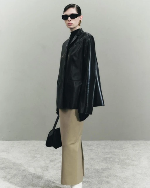 Fashionable woman posing in a black leather jacket and beige skirt, accessorized with sunglasses and a black purse in a studio setting.