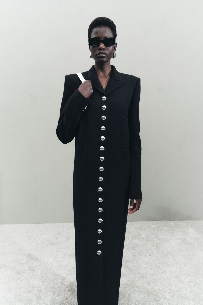 An African American woman poses in a long black coat with silver buttons, wearing sunglasses and carrying a white purse indoors.